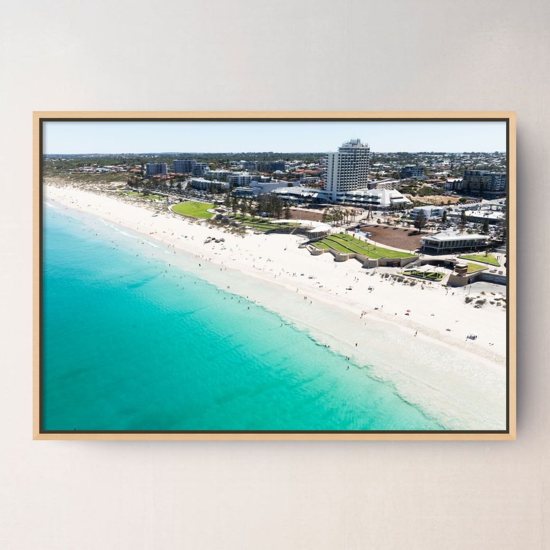 A framed aerial photograph of a Scarborough Beach Perth WA with turquoise ocean water, white sand, and coastal buildings, displayed on a light beige wall.