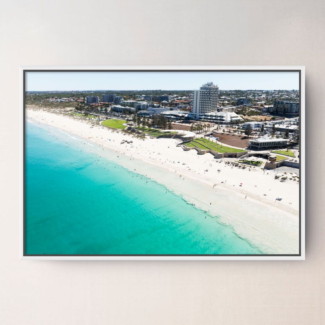 A white framed aerial photograph of a Scarborough Beach Perth WA with turquoise ocean water, white sand, and coastal buildings, displayed on a light beige wall.